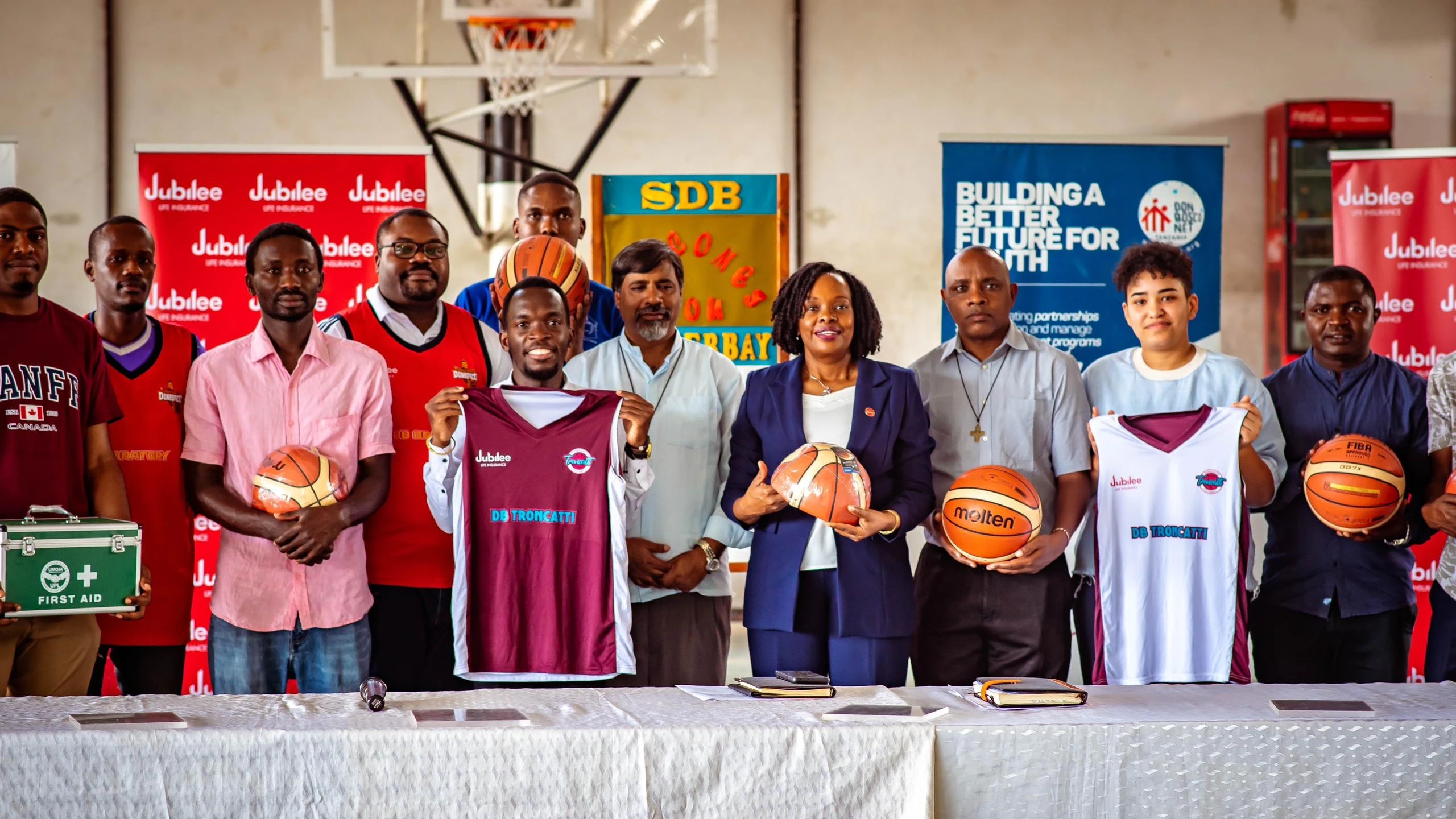 Jubilee Life Insurance Corporation of Tanzania Limited CEO Helena Mzena (4th R), Don Bosco Troncatti captain Nadia Samir (2nd R), Don Bosco basketball teams patron Father Joachim Sivali, (4th L) accompanied by some players and officials showcasing items.